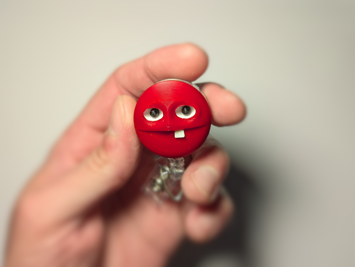 A hand holding a small red novelty lollipop with a cartoon face, featuring large eyes and a single white tooth.
