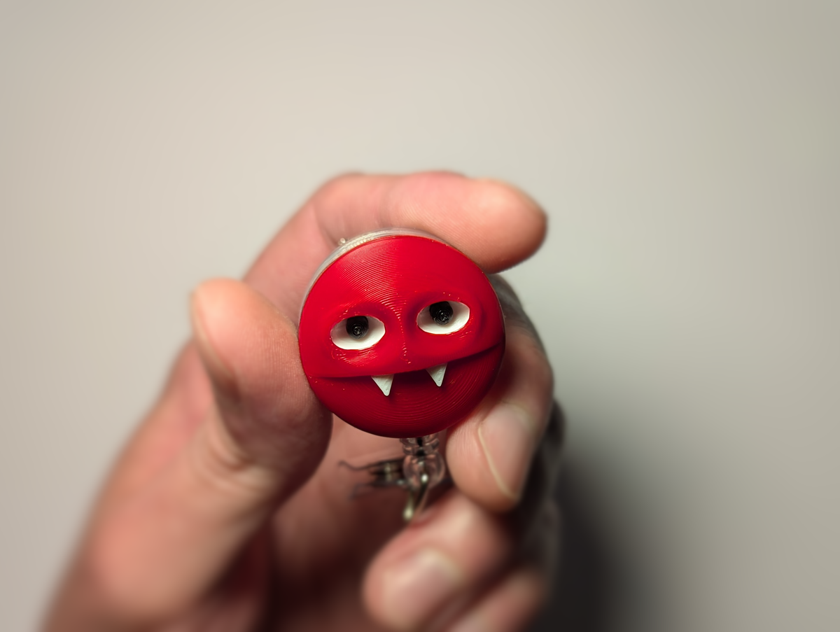 A hand holding a small round red keychain or badge holder with a cartoonish face, featuring large white eyes and two small white fangs, against a plain light gray background.